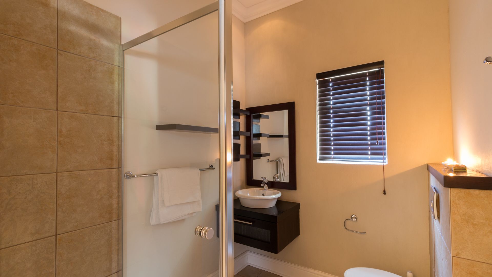 A modern bathroom with a glass shower door, a white sink, a dark vanity, and a window with blinds.