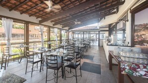 A spacious dining area with multiple tables and chairs, a ceiling fan, and large windows offering views of the outside. - Hotel Ilha Branca Inn (Búzios)