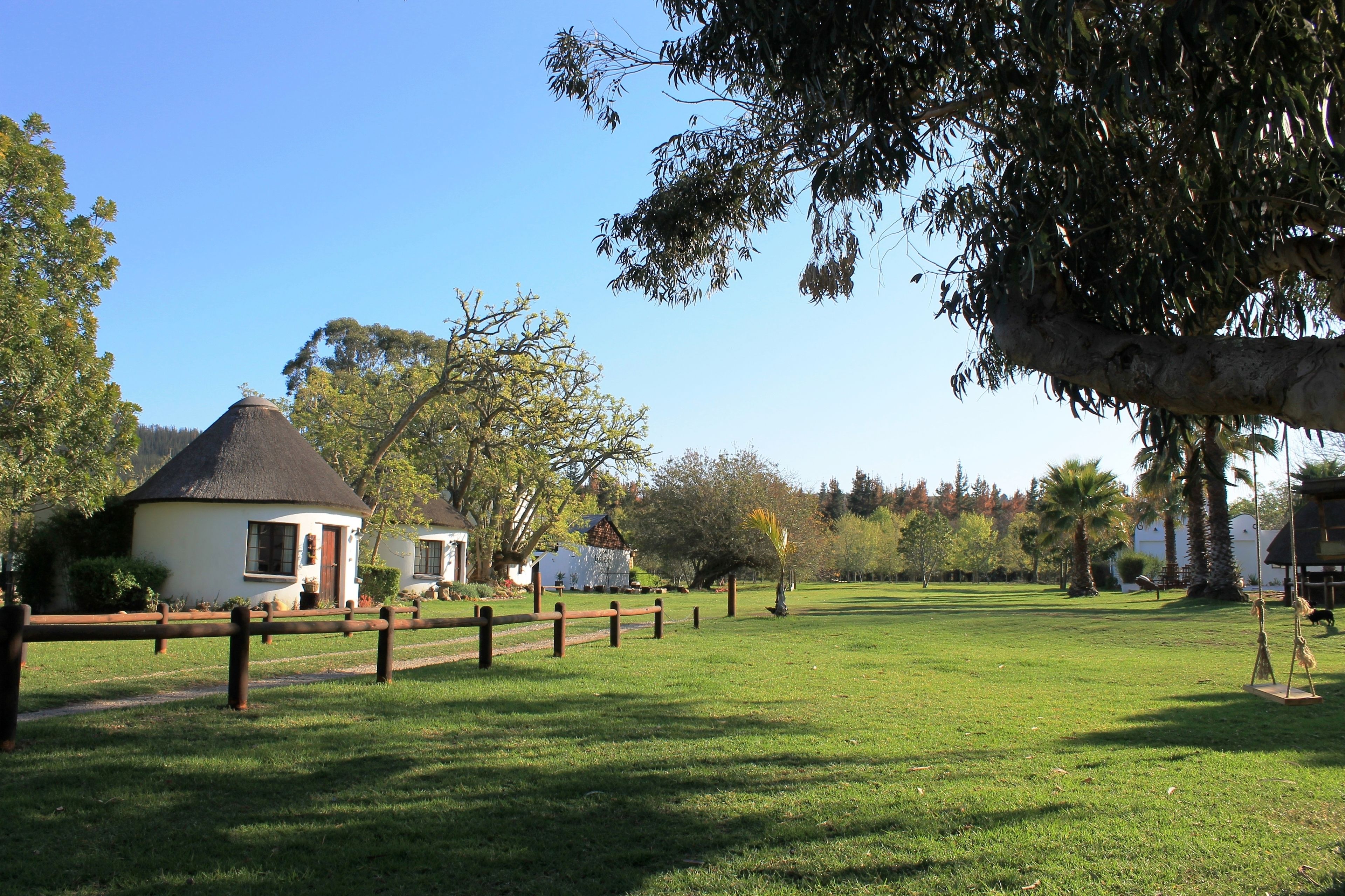 A grassy area with a small white building with a thatched roof, surrounded by trees and a wooden fence.