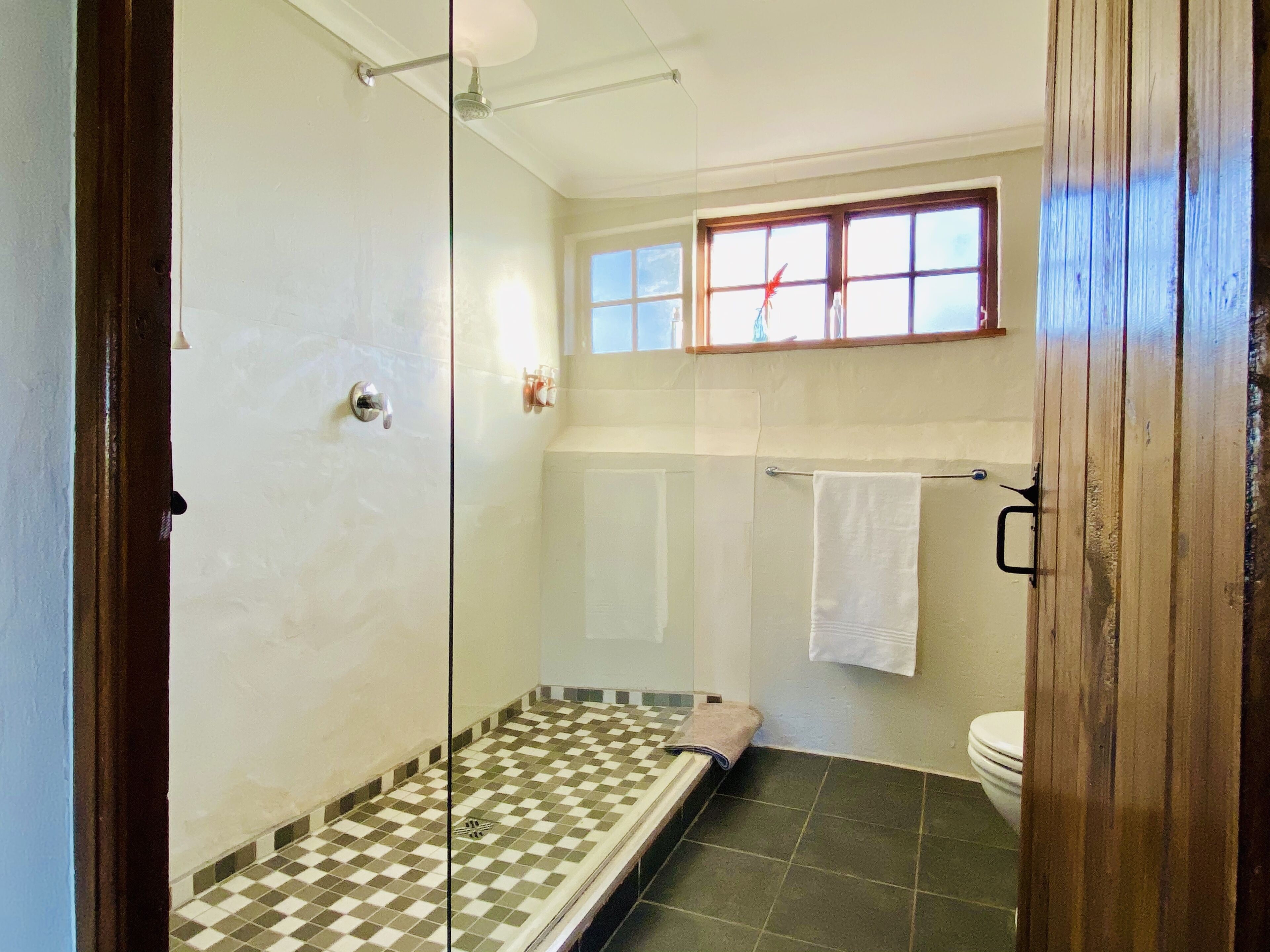 A bathroom with a checkered floor, a shower, a toilet, and a window above the sink.