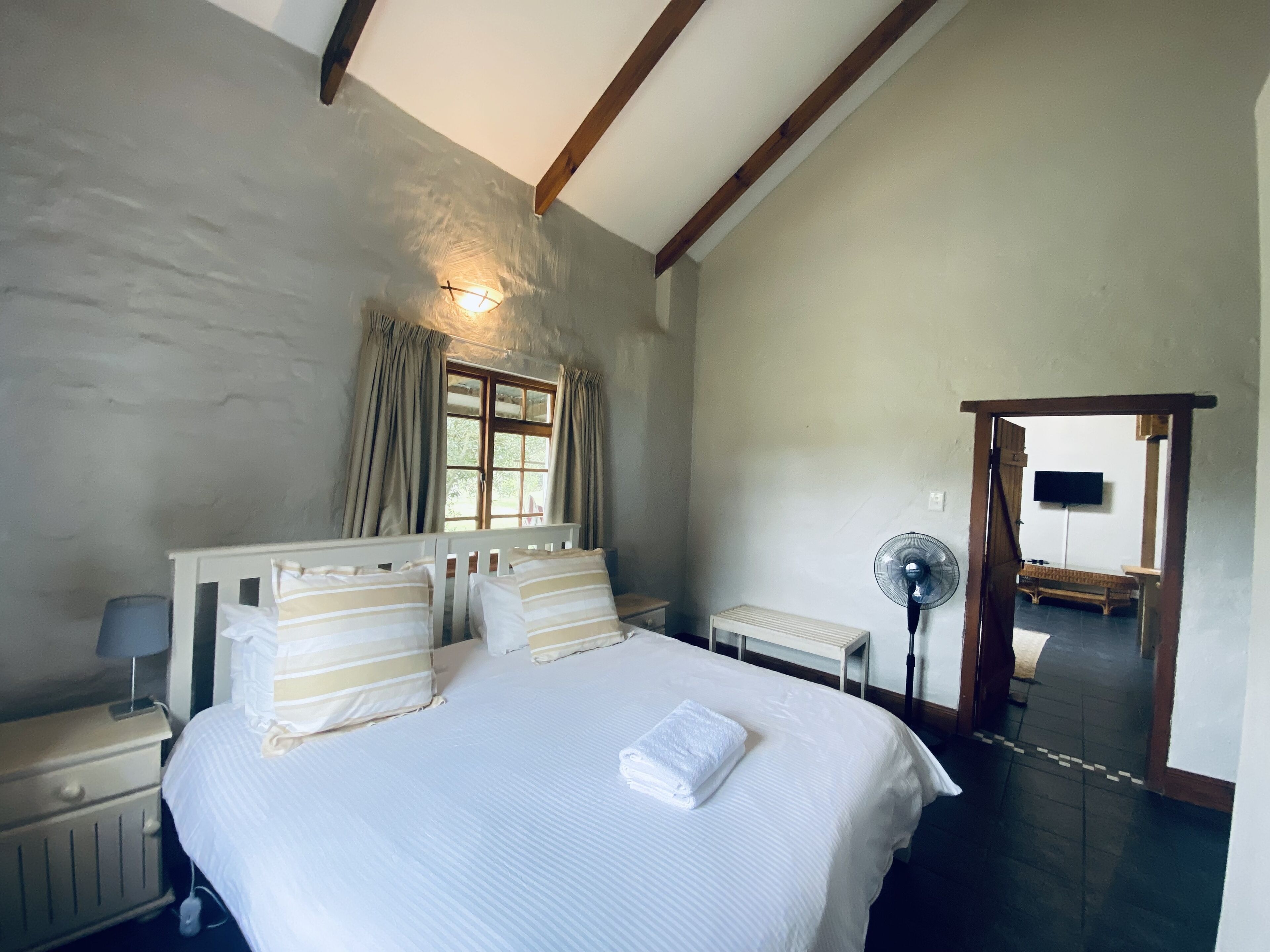 Un dormitorio con cama, mesitas de noche, un ventilador y una ventana con cortinas.