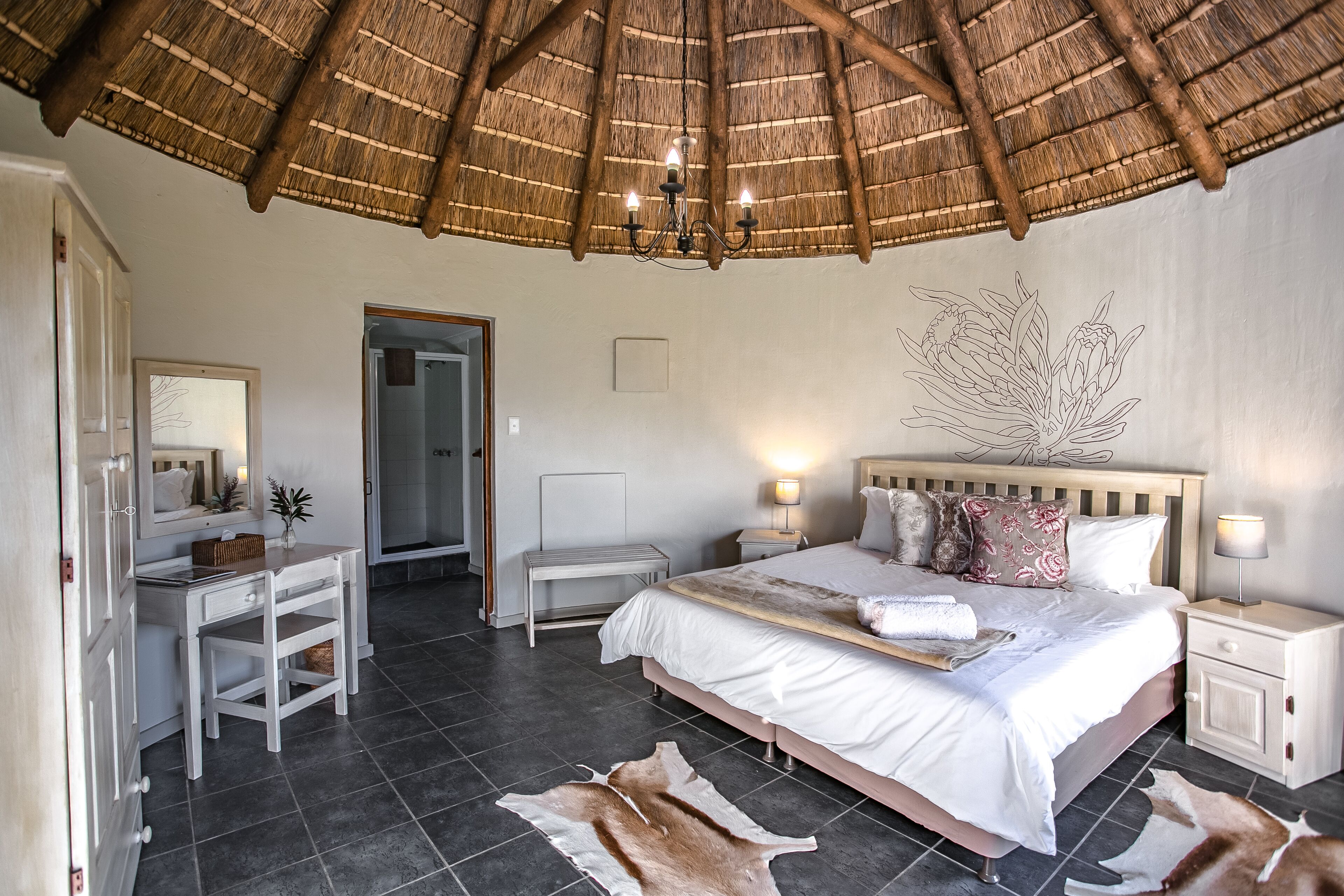 A bedroom with a bed, bedside tables, a desk, a chair, a mirror, a thatched roof, and a wall art piece.