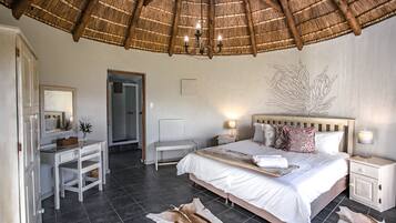 A bedroom with a bed, bedside tables, a desk, a chair, a mirror, a thatched roof, and a wall art piece.