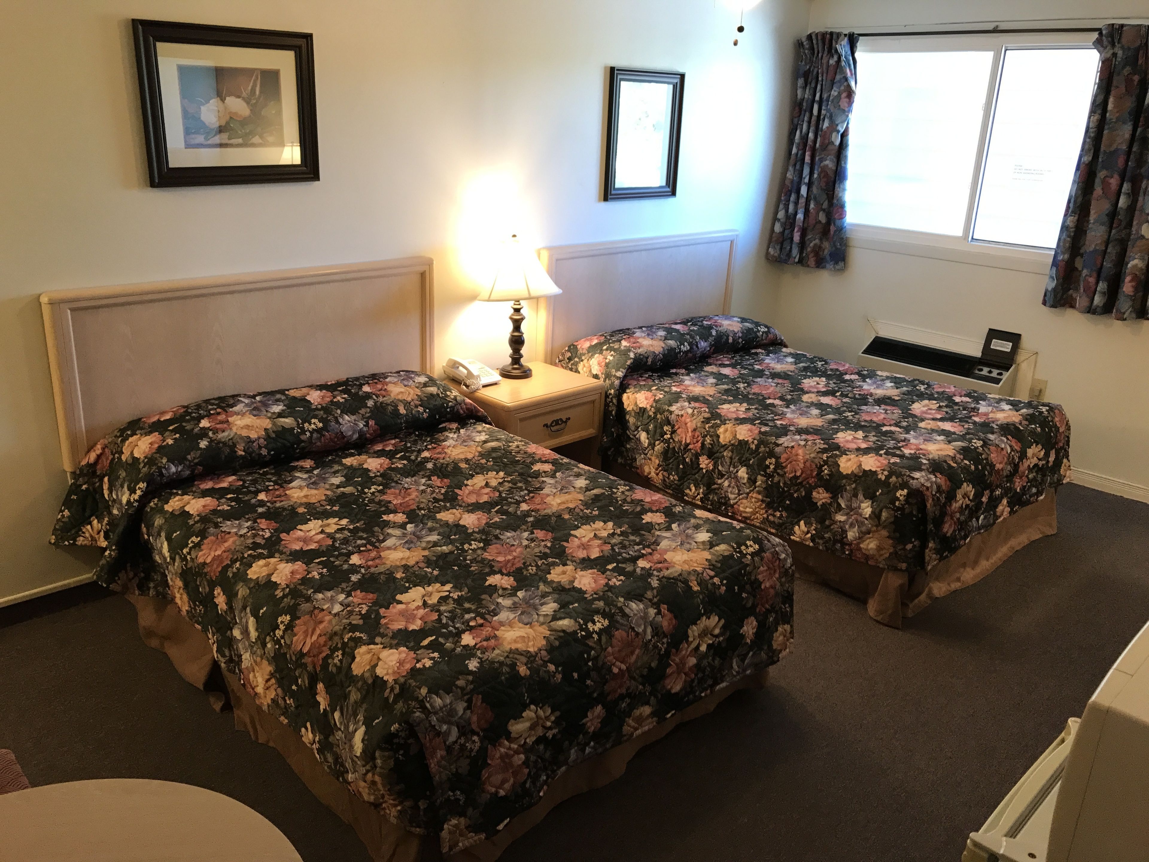 A hotel room with two beds, a bedside table with a lamp, a telephone, and a window with curtains.