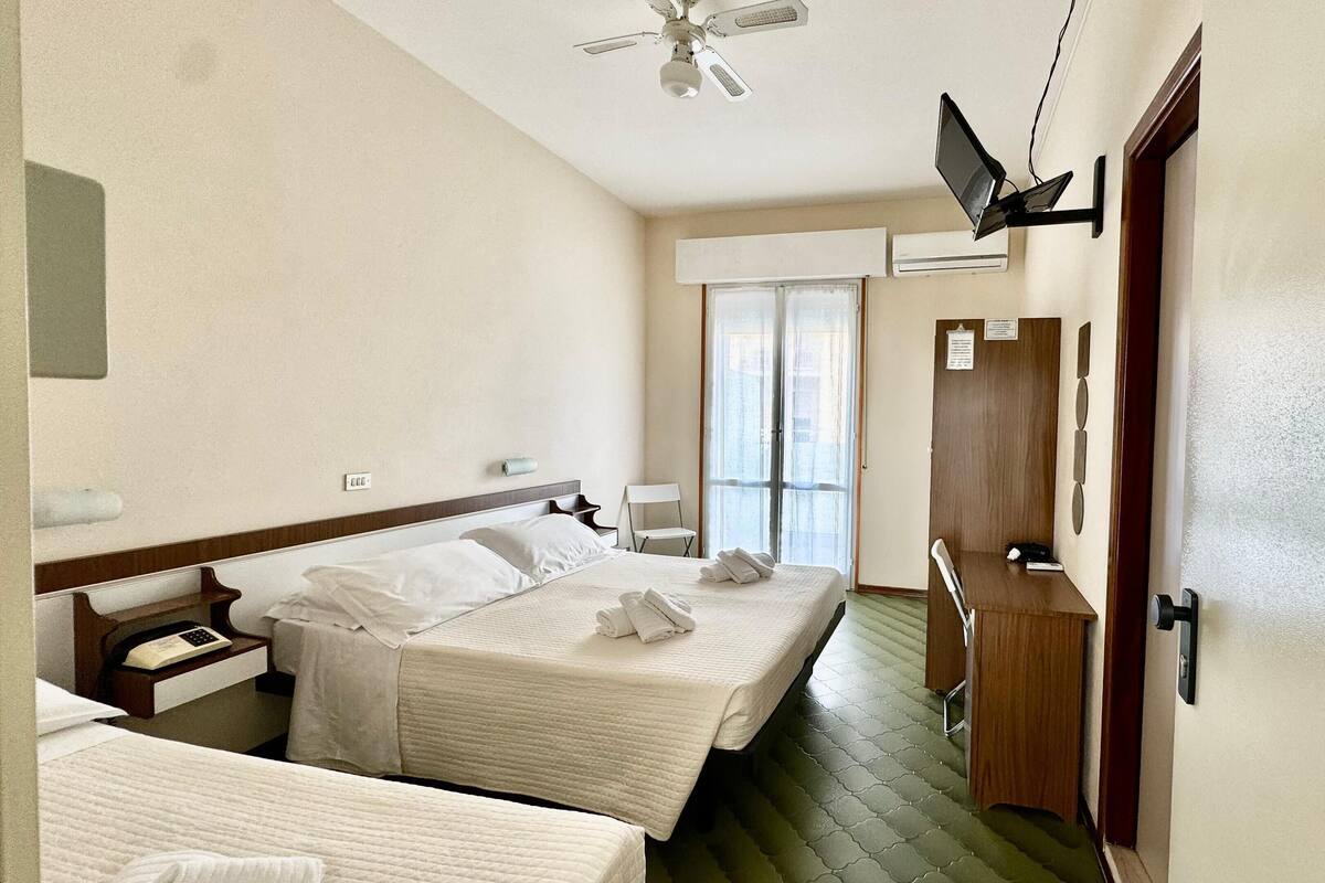 Une chambre d’hôtel avec deux lits, un ventilateur de plafond, un appareil de climatisation, un téléviseur fixé au mur et une porte coulissante donnant sur un balcon.