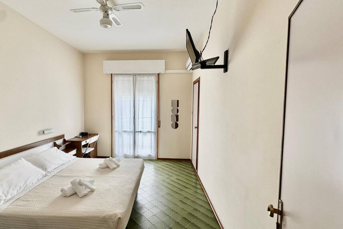 Une chambre d’hôtel équipée d’un lit, d’un bureau, d’une chaise, d’un ventilateur de plafond et d’une fenêtre avec des rideaux.