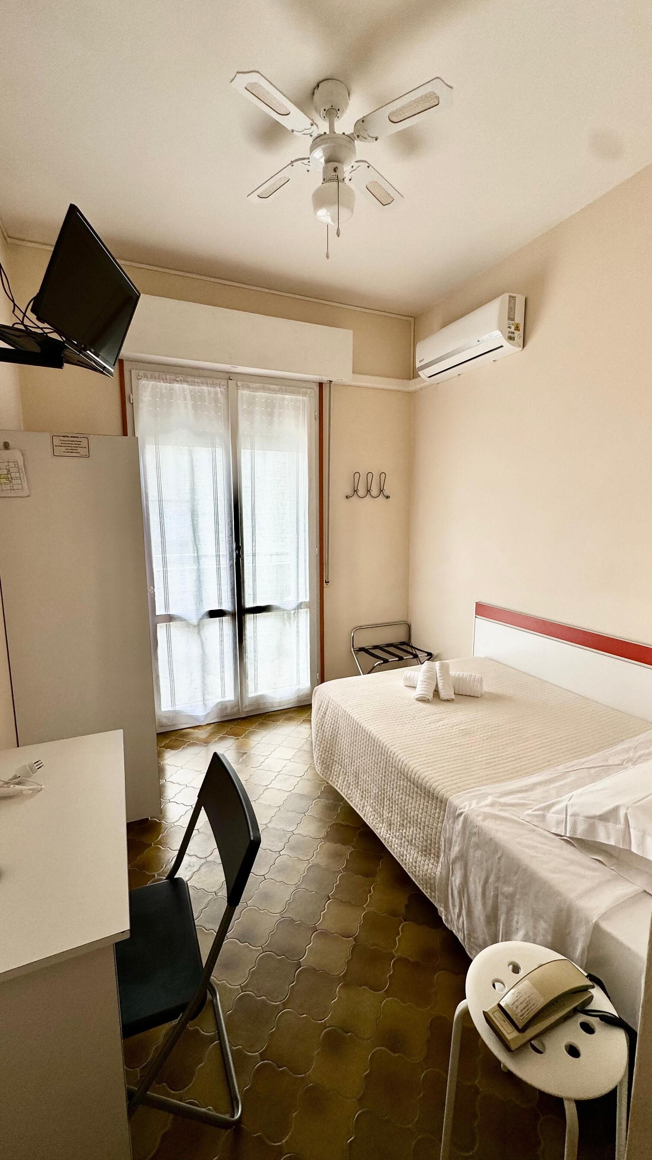 Une chambre d’hôtel équipée d’un lit, d’un bureau, d’une chaise, d’un ventilateur de plafond, d’un appareil de climatisation et d’une fenêtre avec des rideaux.