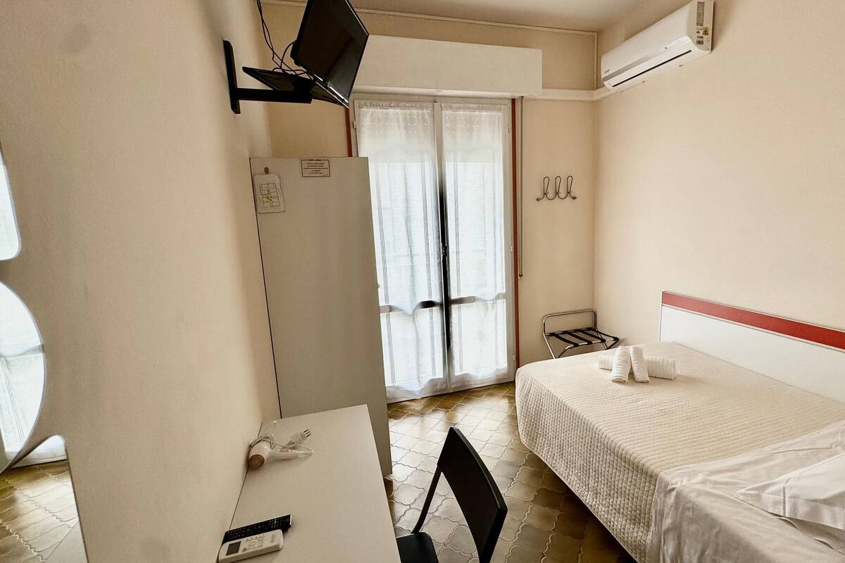 Une chambre d’hôtel équipée d’un lit, d’un bureau, d’une chaise, d’un miroir, d’une fenêtre avec des rideaux et d’un appareil de climatisation.