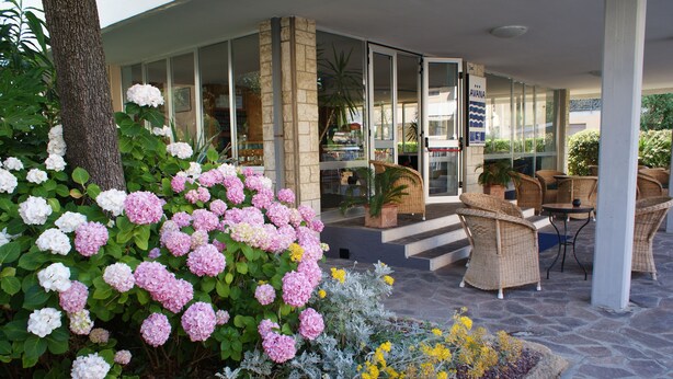L’entrée d’un hôtel s’ouvre sur un patio abrité, aménagé avec des meubles en osier et agrémenté de diverses plantes, notamment des hortensias et d’autres arbustes à fleurs.