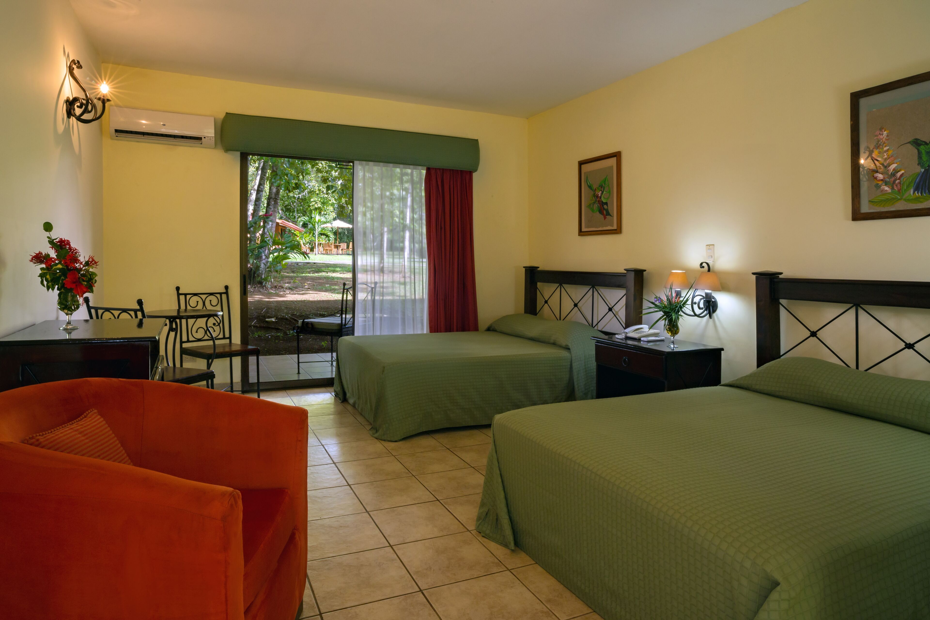 Habitación de hotel con dos camas, una mesa de comedor, una silla y ventana con vista a follaje.