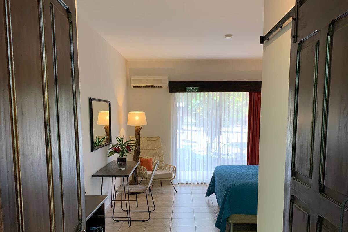Habitación de hotel con cama, escritorio con silla, mesita de noche con lámpara y ventana con cortinas.