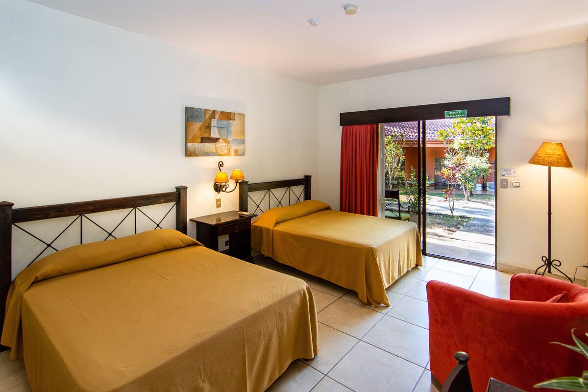 Habitación de hotel con dos camas, una mesita de noche, una lámpara, una silla roja y vistas al exterior.