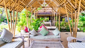 A bamboo pavilion with a thatched roof, surrounded by lush greenery and outdoor seating with cushions.