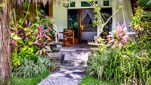 A tropical garden entrance with a thatched roof, a stone pathway, and a bed visible inside.