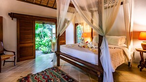 A four-poster bed with a canopy, a wooden chair, a patterned rug, and a view of lush greenery outside.