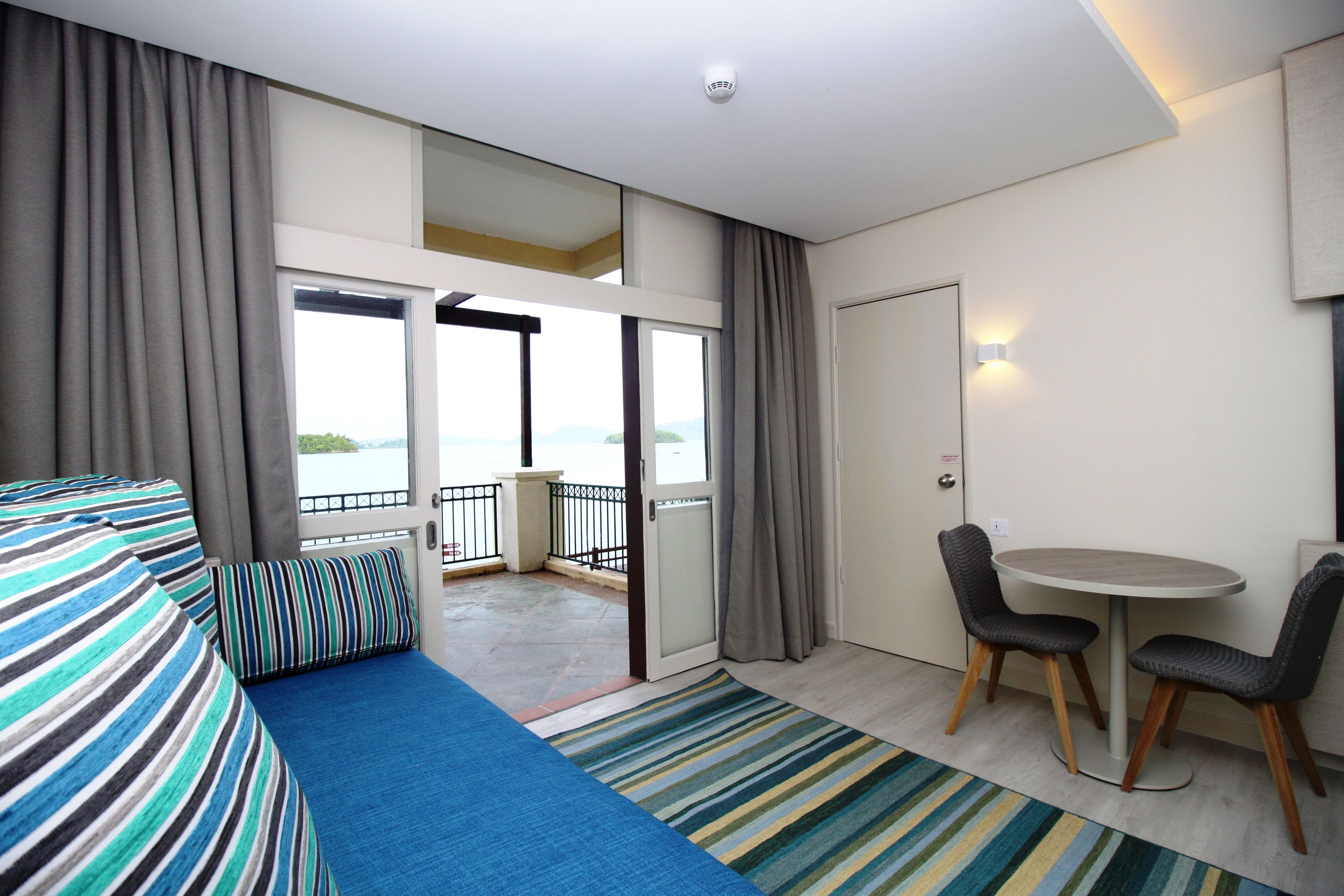 A hotel room with a balcony, a sofa, a dining table with chairs, and a view of the sea.