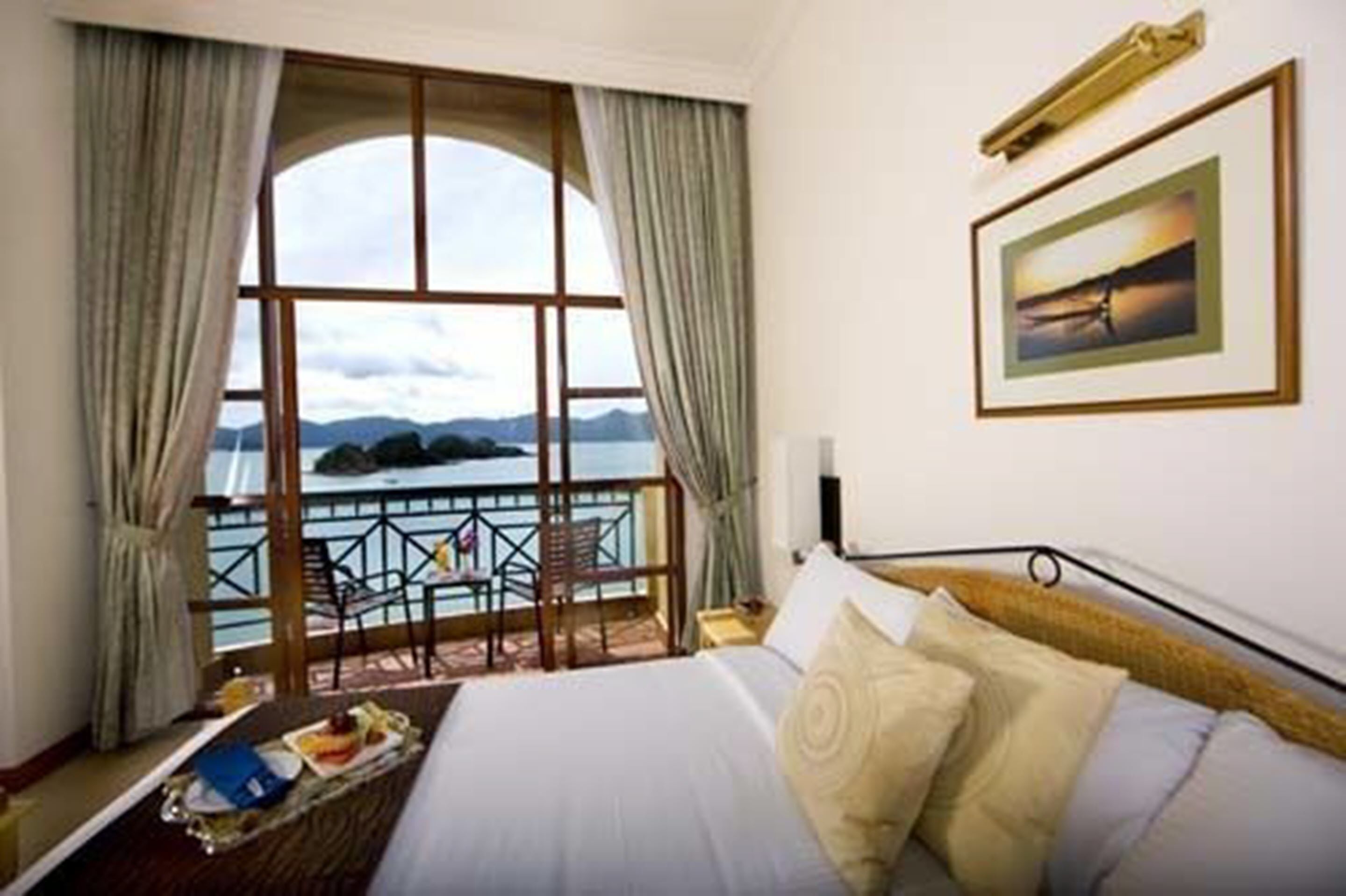 Habitación de hotel con un ventanal que ofrece vistas al mar y a las montañas. Cuenta con una cama con ropa blanca y un cuadro en la pared.