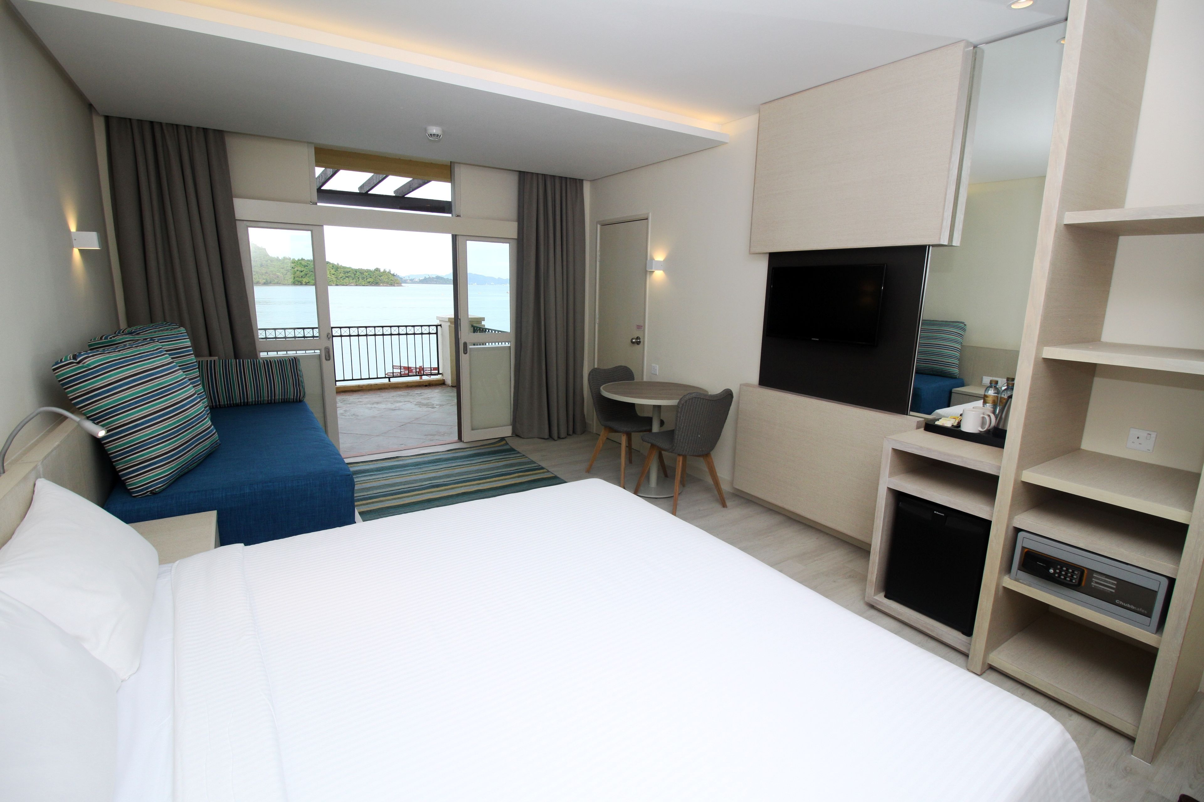 A hotel room with a bed, a desk, a TV, and a balcony with a view of the water.
