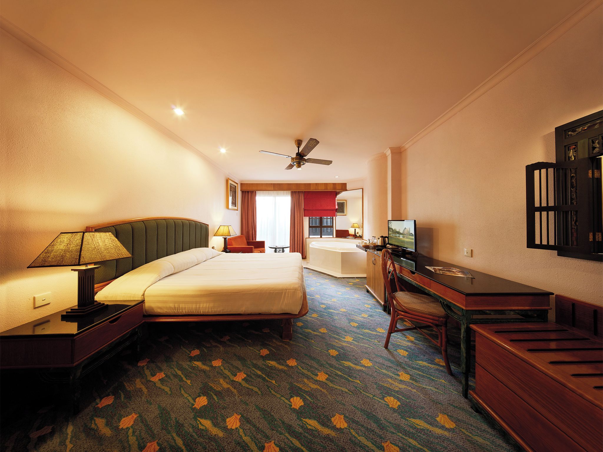 A hotel room with a bed, a desk with a computer, a chair, a TV, a ceiling fan, and a bathroom visible through an open door.