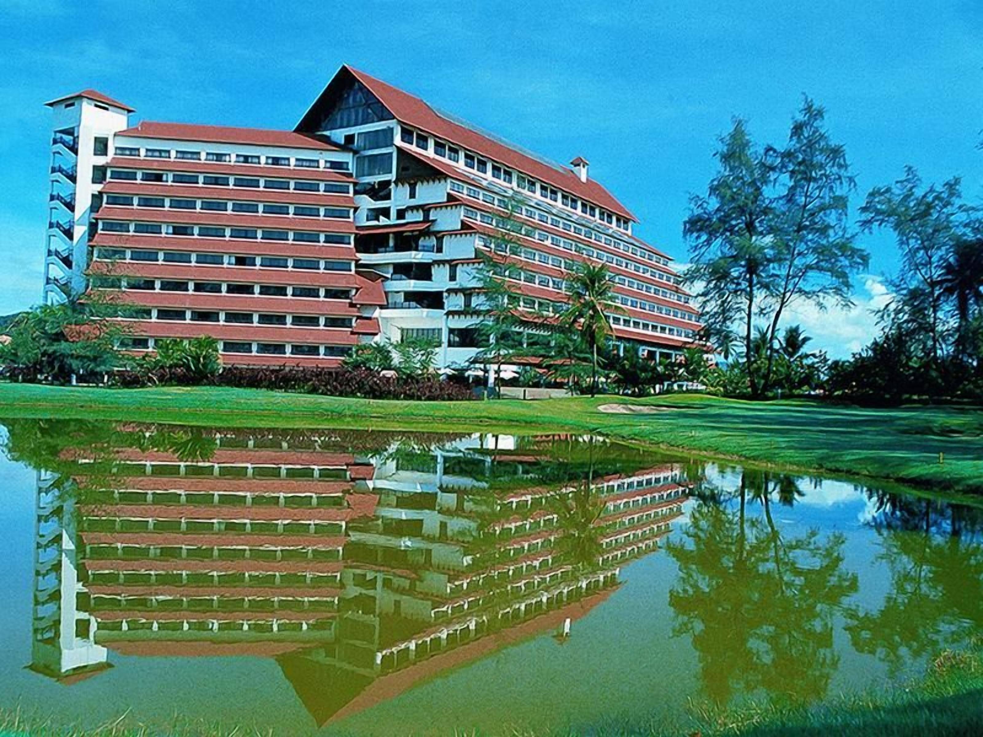 A multi-story hotel with a reflection in a pond, surrounded by greenery and palm trees.