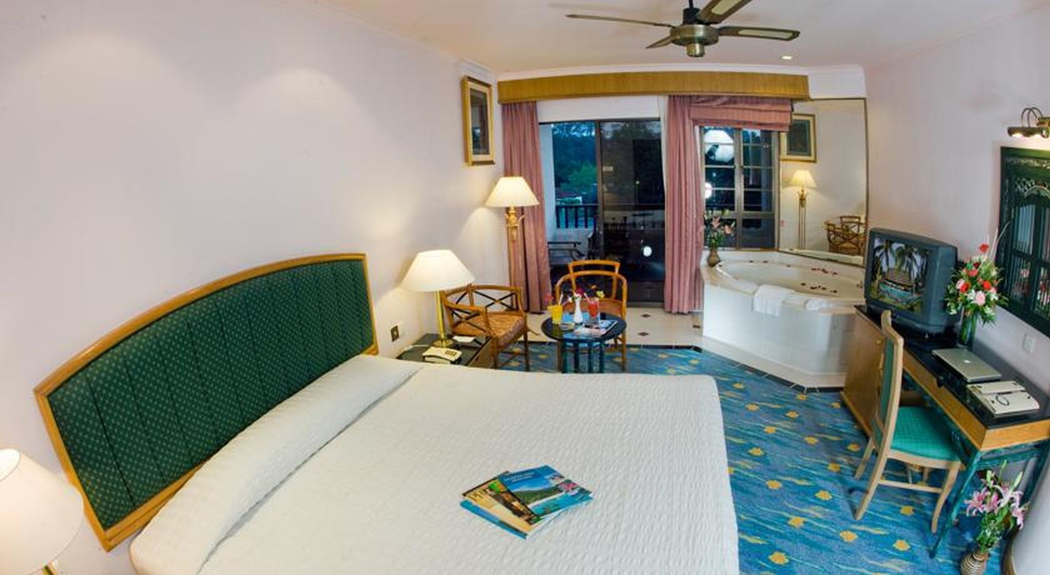 A hotel room with a bed, a desk with a computer, a TV, a chair, a small table with drinks, and a balcony with a view.