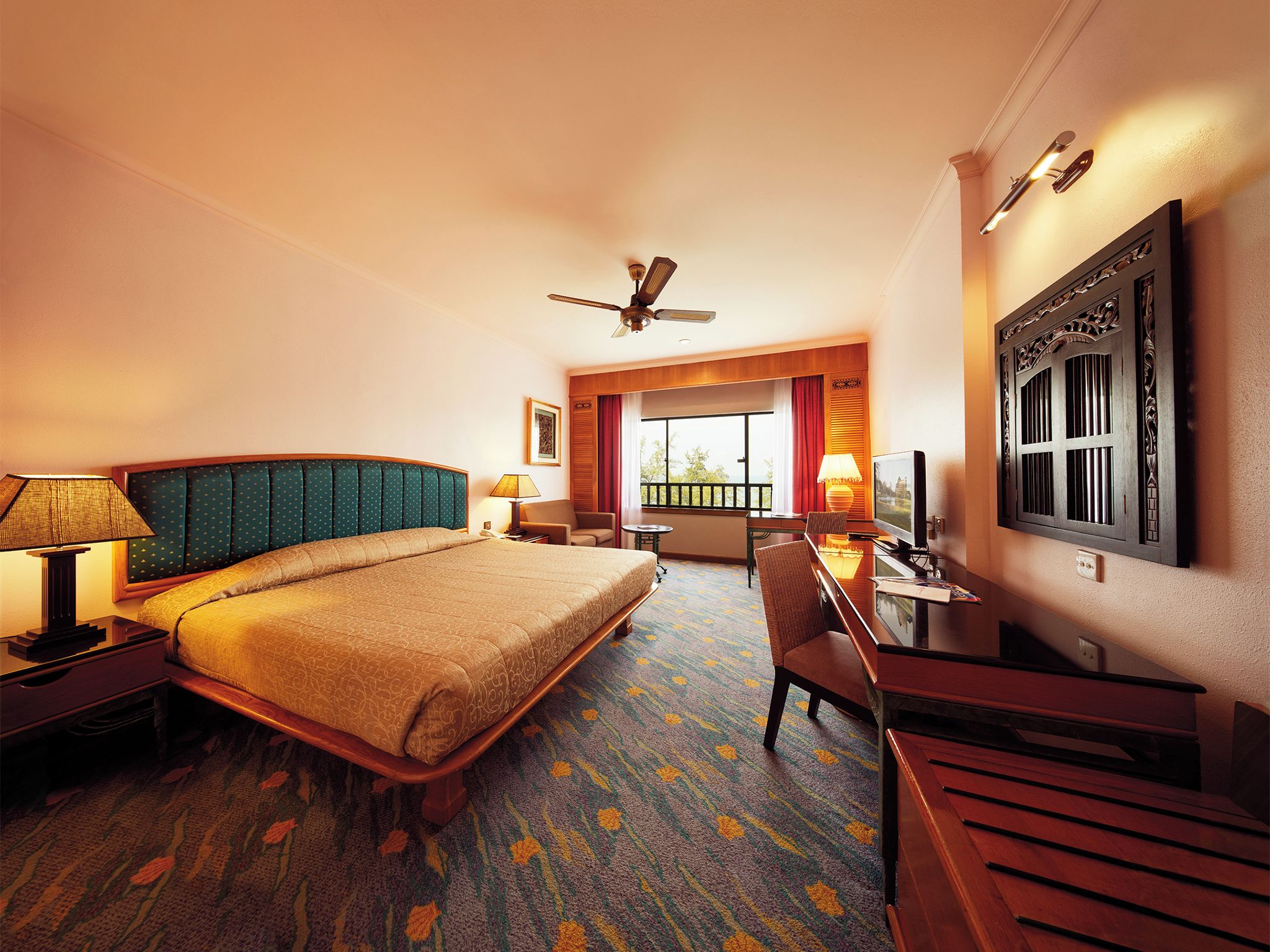 A hotel room with a large bed, a desk with a chair, a television, a ceiling fan, and a window with a view of the outdoors.