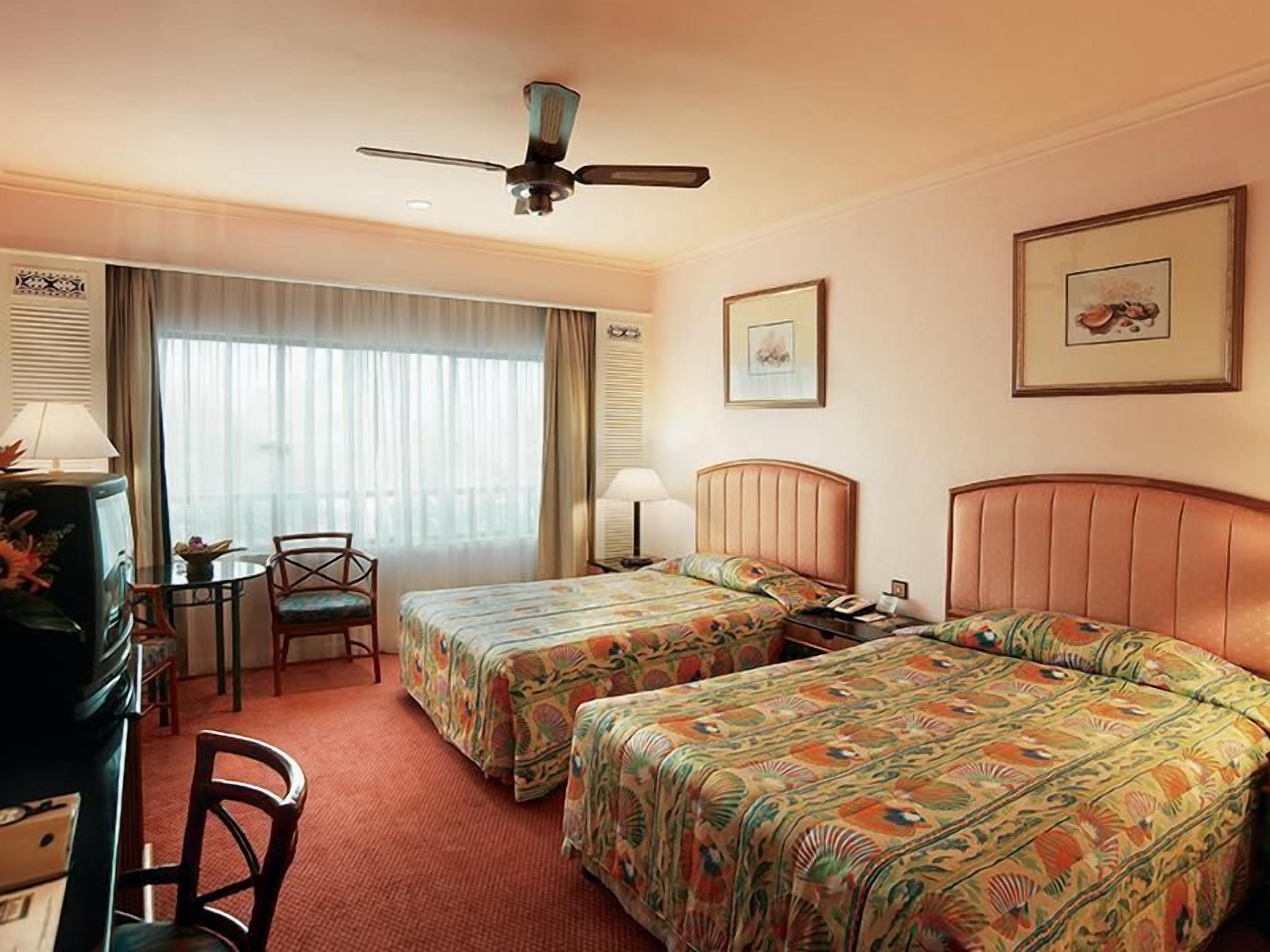 A hotel room with two beds, a desk, a chair, a ceiling fan, and a window with curtains.