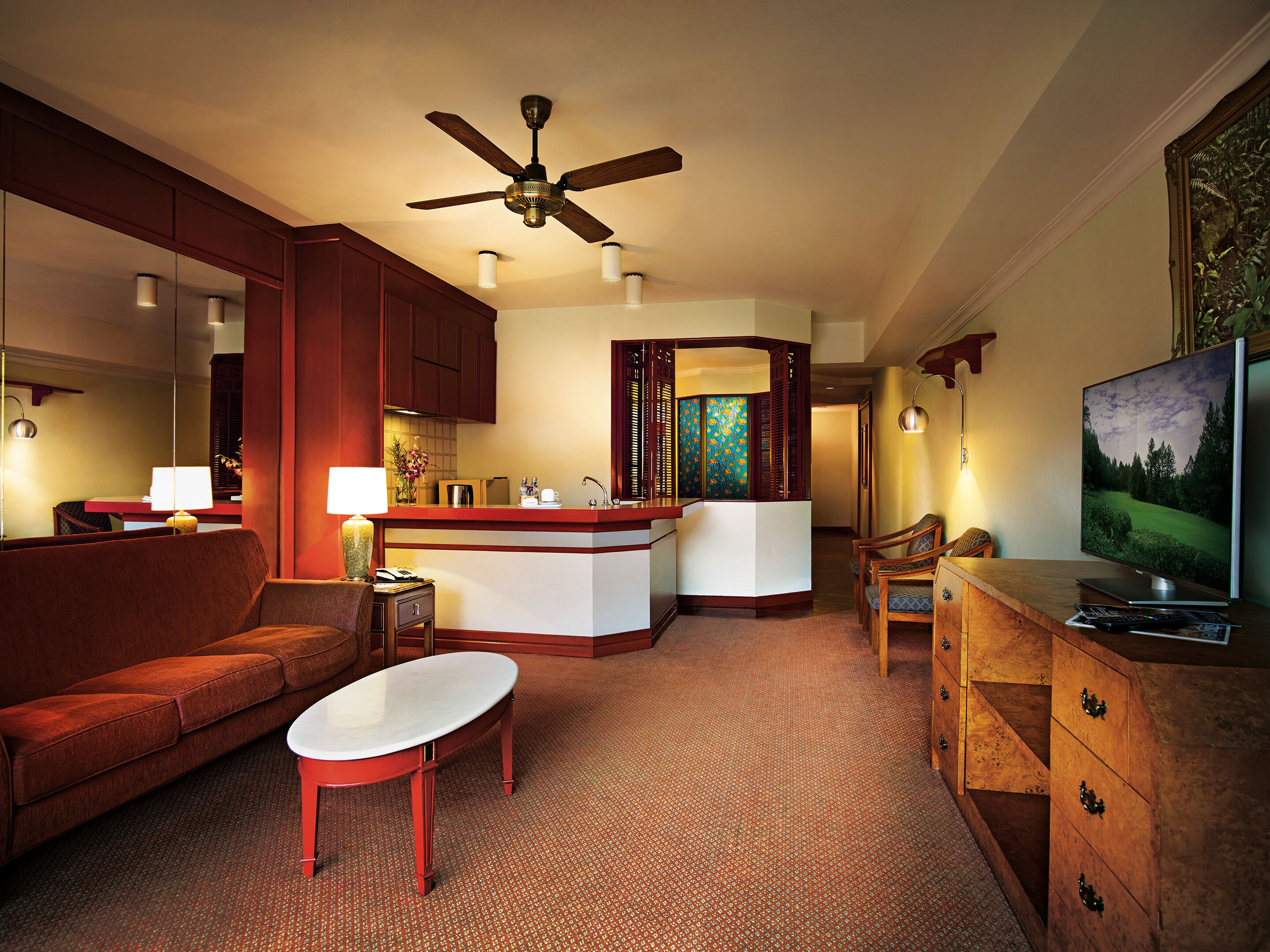 A hotel room with a living area, a kitchenette, a ceiling fan, a TV, and a red carpet.