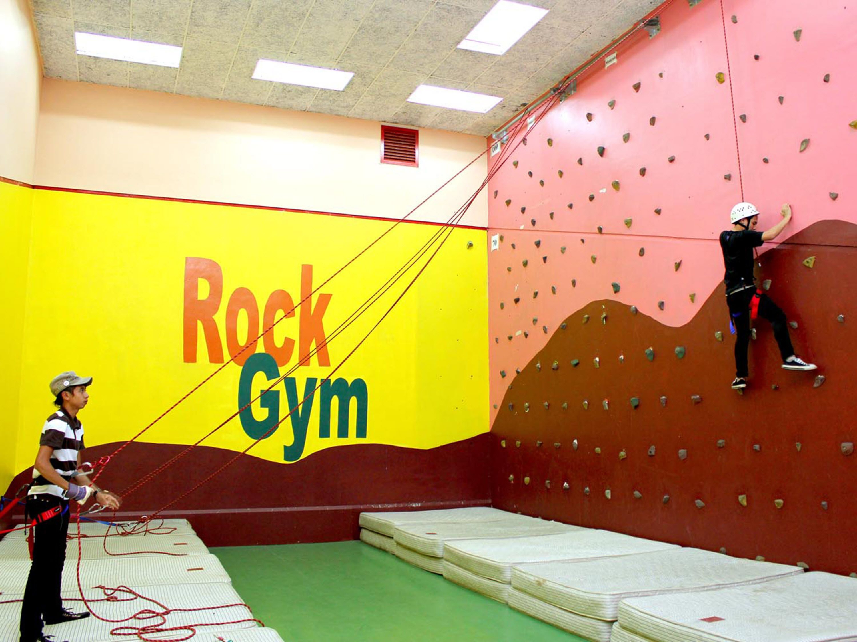 Een klimhal met een grote wand waarop “Rock Gym” staat en getopte matten op de vloer.