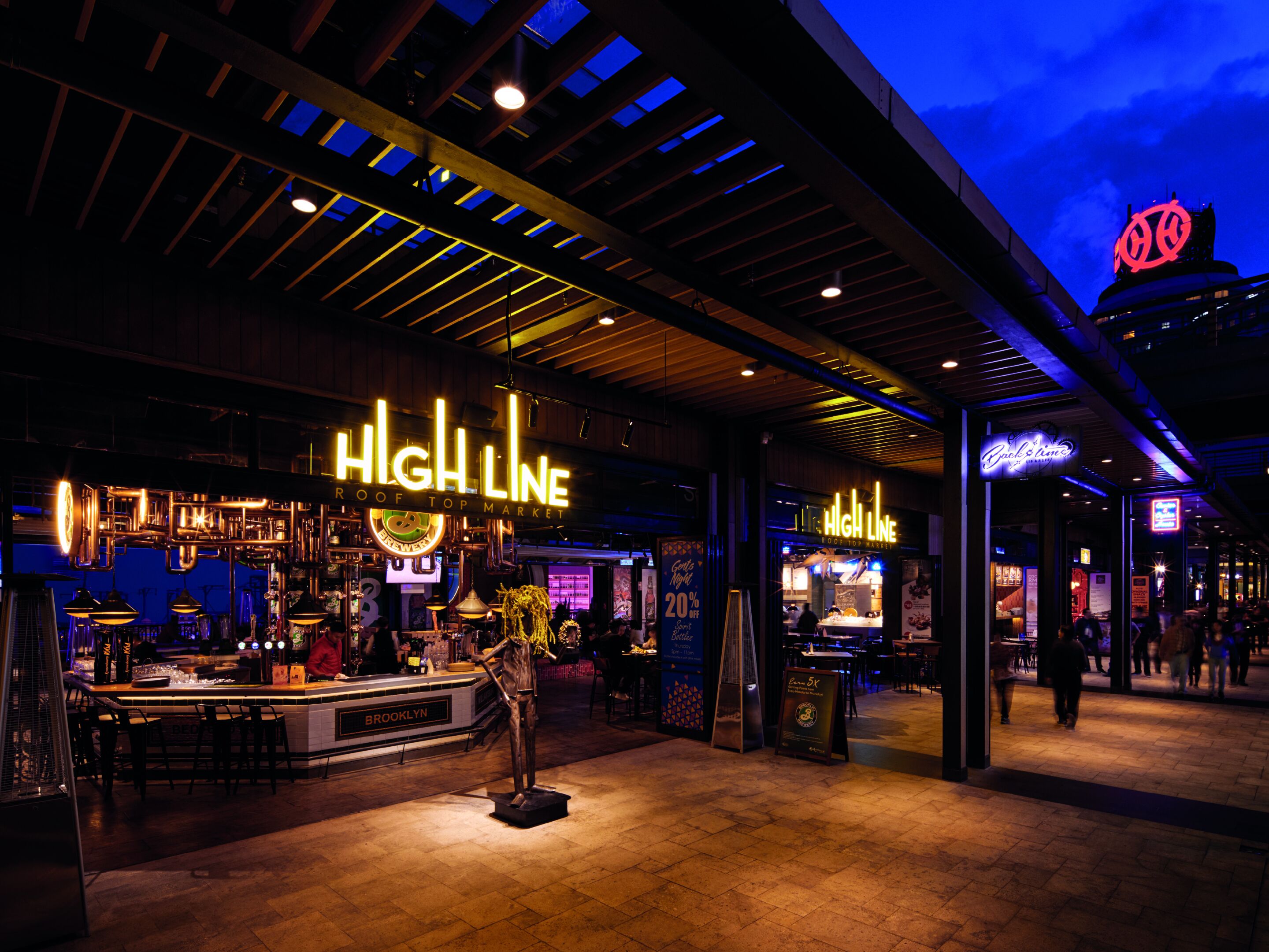 A rooftop bar with a neon sign, outdoor seating, and a view of the city skyline at dusk.