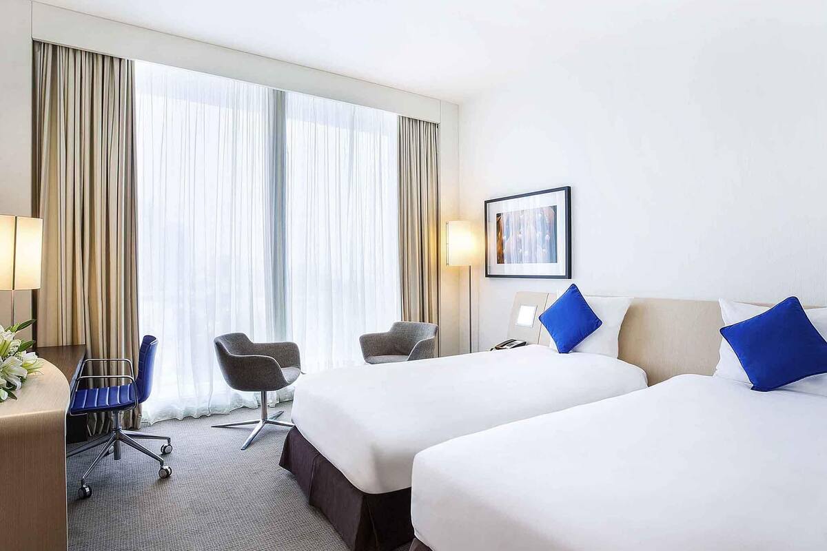 Een hotelkamer met twee bedden, een bureau met een stoel, een lamp en een groot raam met gordijnen.