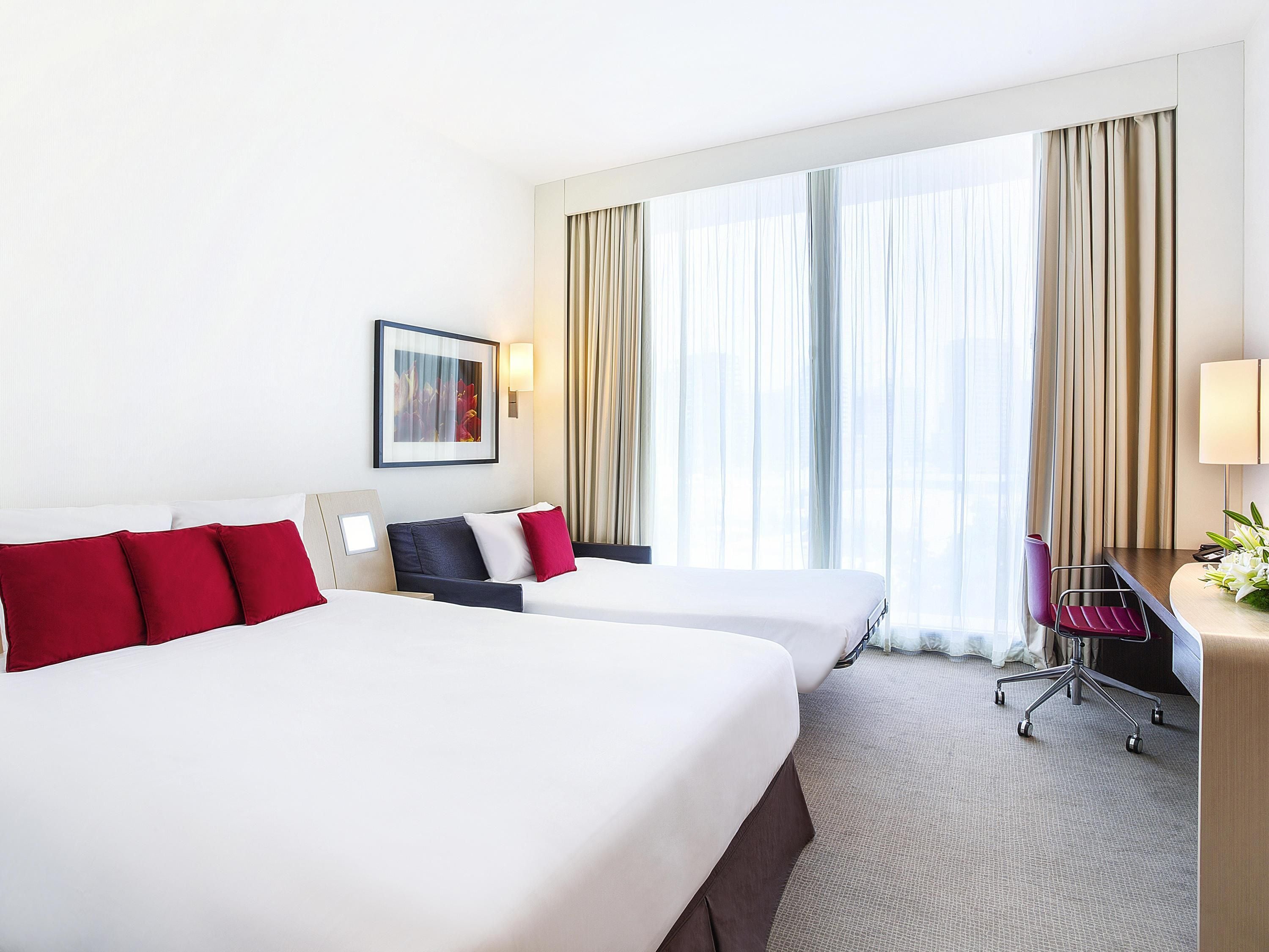 Habitación de hotel con dos camas, un escritorio, una silla y un amplio ventanal con cortinas.