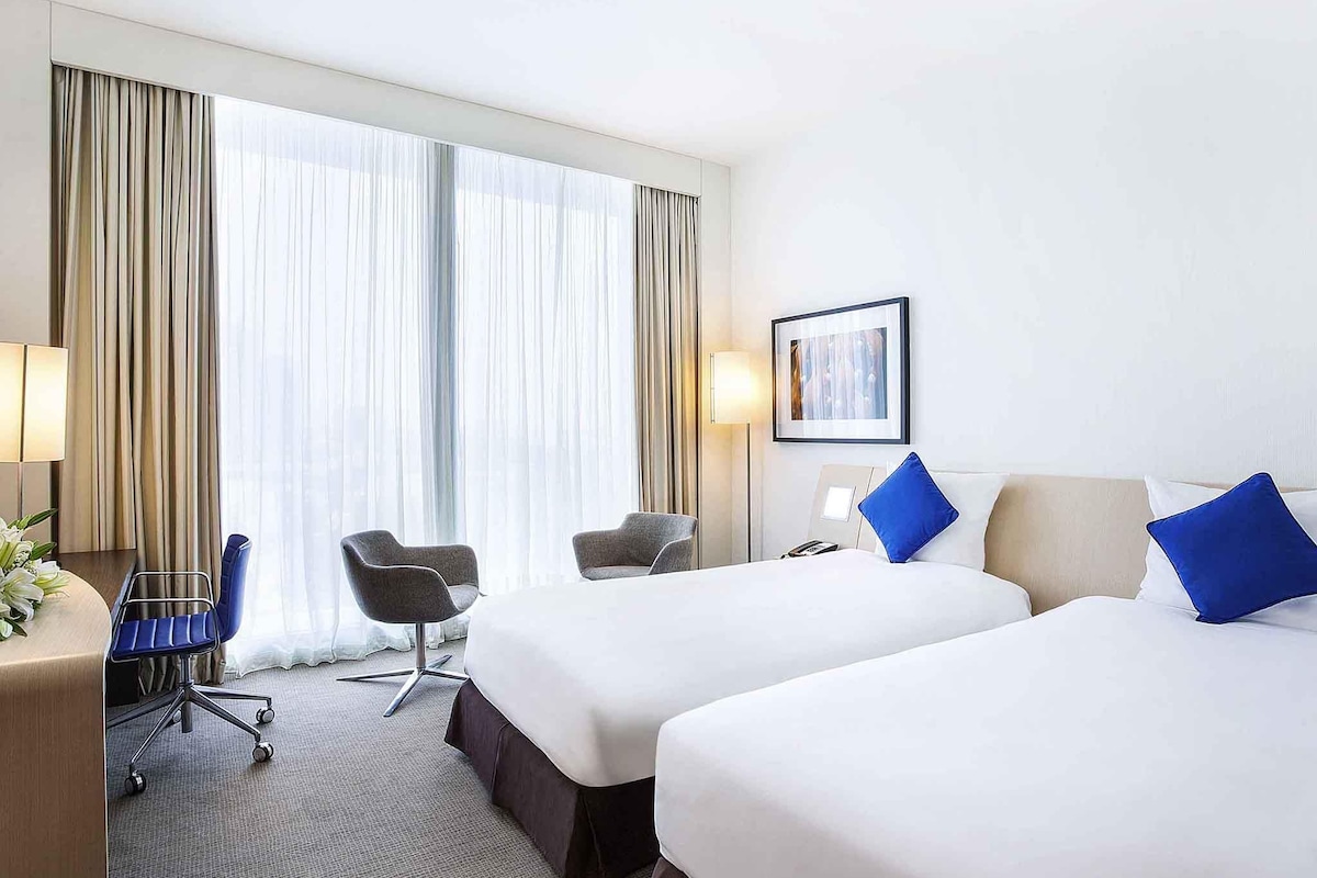 Een hotelkamer met twee bedden, een bureau met een stoel, een lamp en een groot raam met gordijnen.