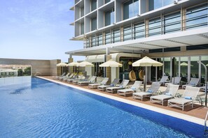 A swimming pool with sun loungers and umbrellas, a modern building with large windows, and a clear sky.