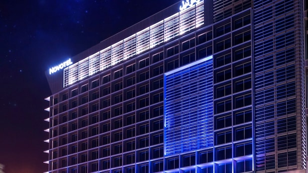 A modern hotel with a prominent 'Novotel' sign, featuring a grid-like facade and illuminated blue lighting.