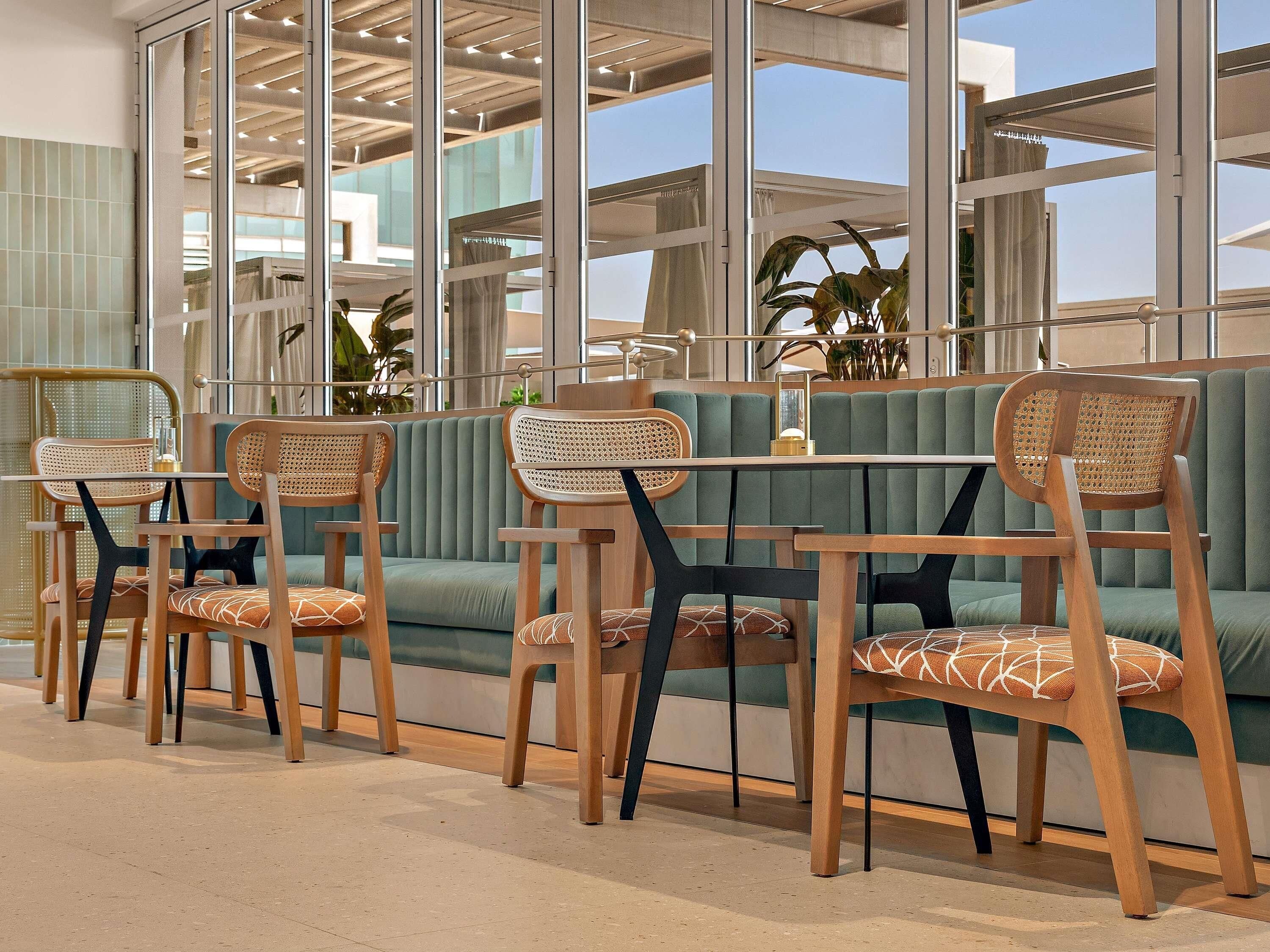A seating area with wooden chairs and a cushioned bench, large windows, and indoor plants.