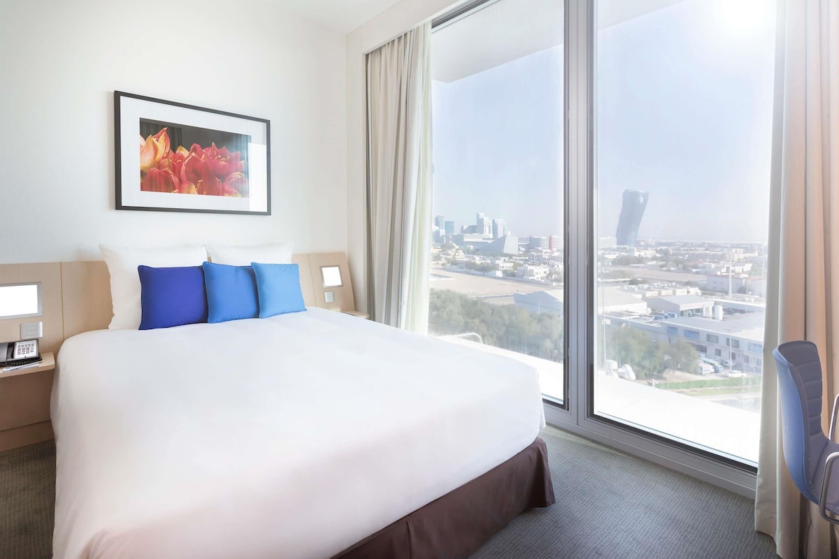 Hotelkamer met een groot bed, stadszicht, een ingelijst schilderij aan de muur en een nachtkastje met een telefoon.