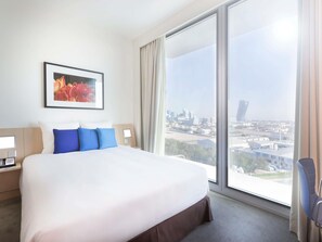 A hotel room with a large bed, a city view, a framed picture on the wall, and a bedside table with a phone.