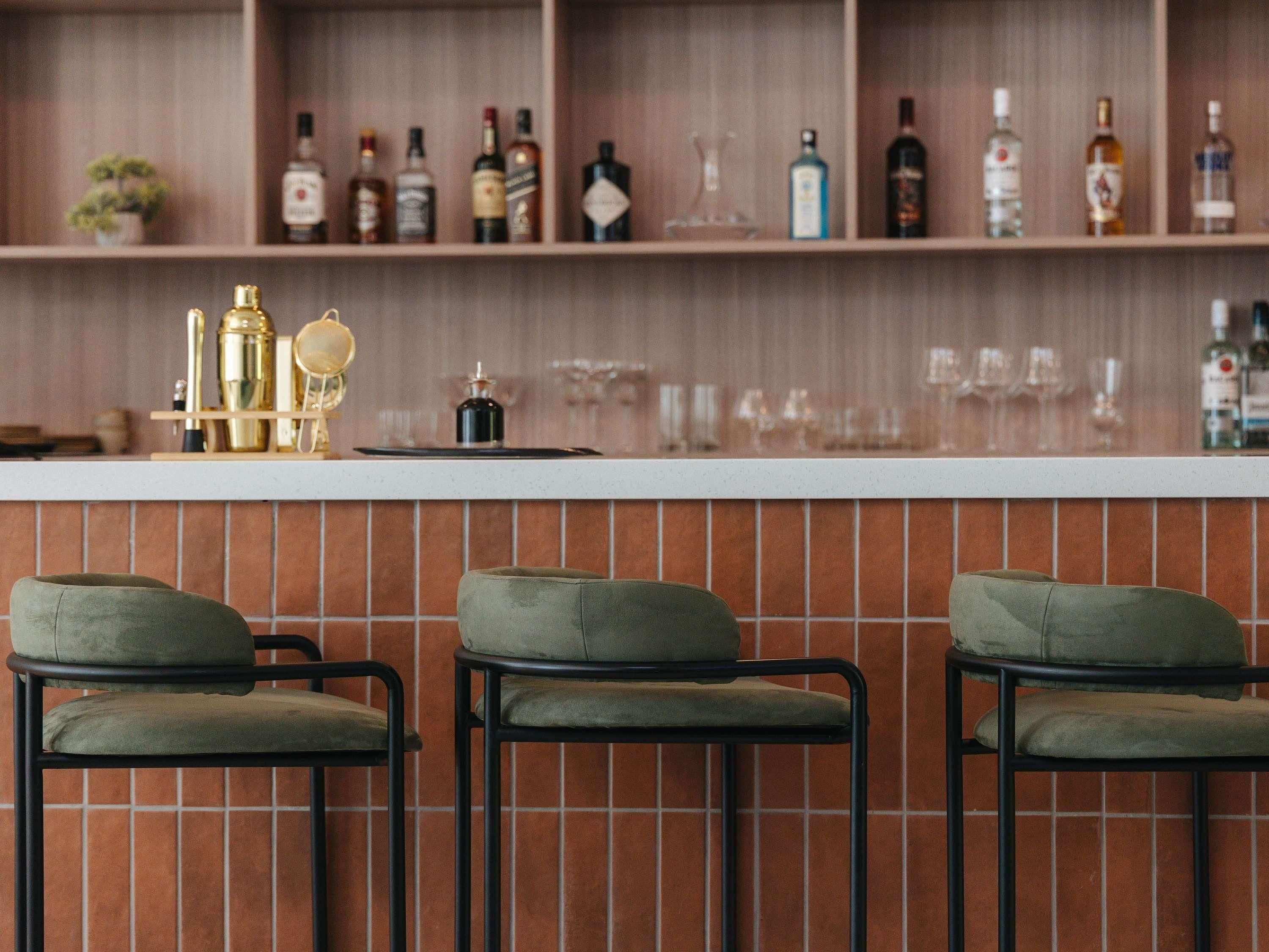 A bar area with three green cushioned bar stools, a white countertop, and shelves with various bottles and glasses.