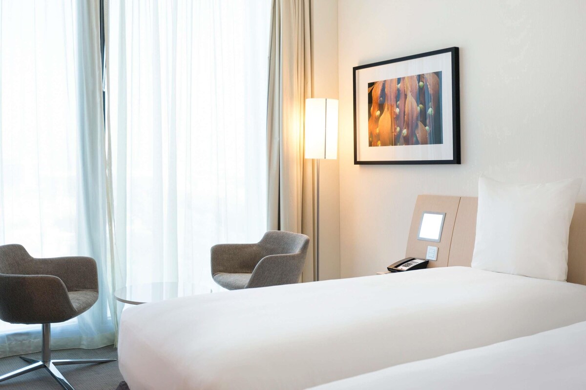 Une chambre d’hôtel avec un lit, deux fauteuils, une table de chevet avec un téléphone, une lampe et un tableau encadré au mur.