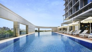 Une piscine à débordement située sur le toit, agrémentée de chaises longues et de parasols, et entourée d’une architecture moderne.
