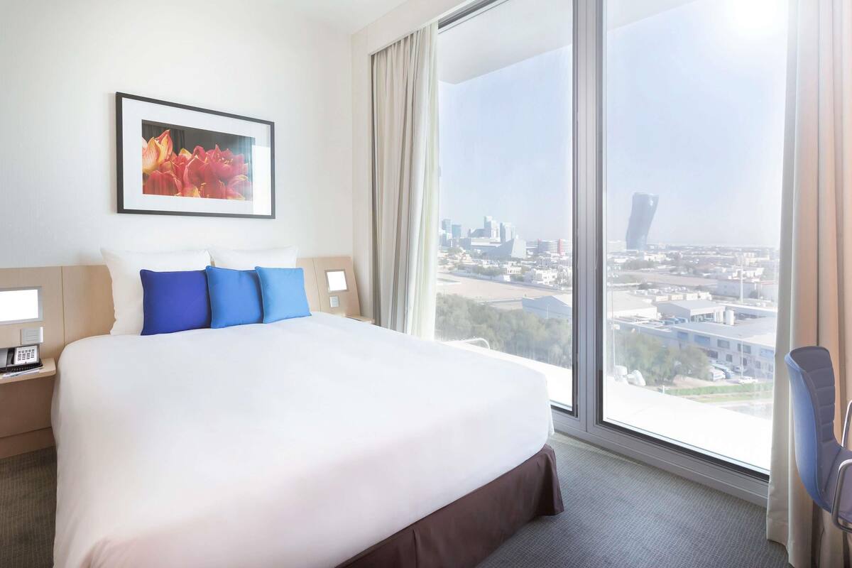 Hotelkamer met een groot bed, stadszicht, een ingelijst schilderij aan de muur en een nachtkastje met een telefoon.