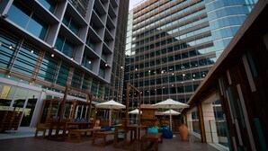 Een moderne binnentuin met houten zitplaatsen en parasols, omgeven door hoge gebouwen met reflecterende glazen ramen.