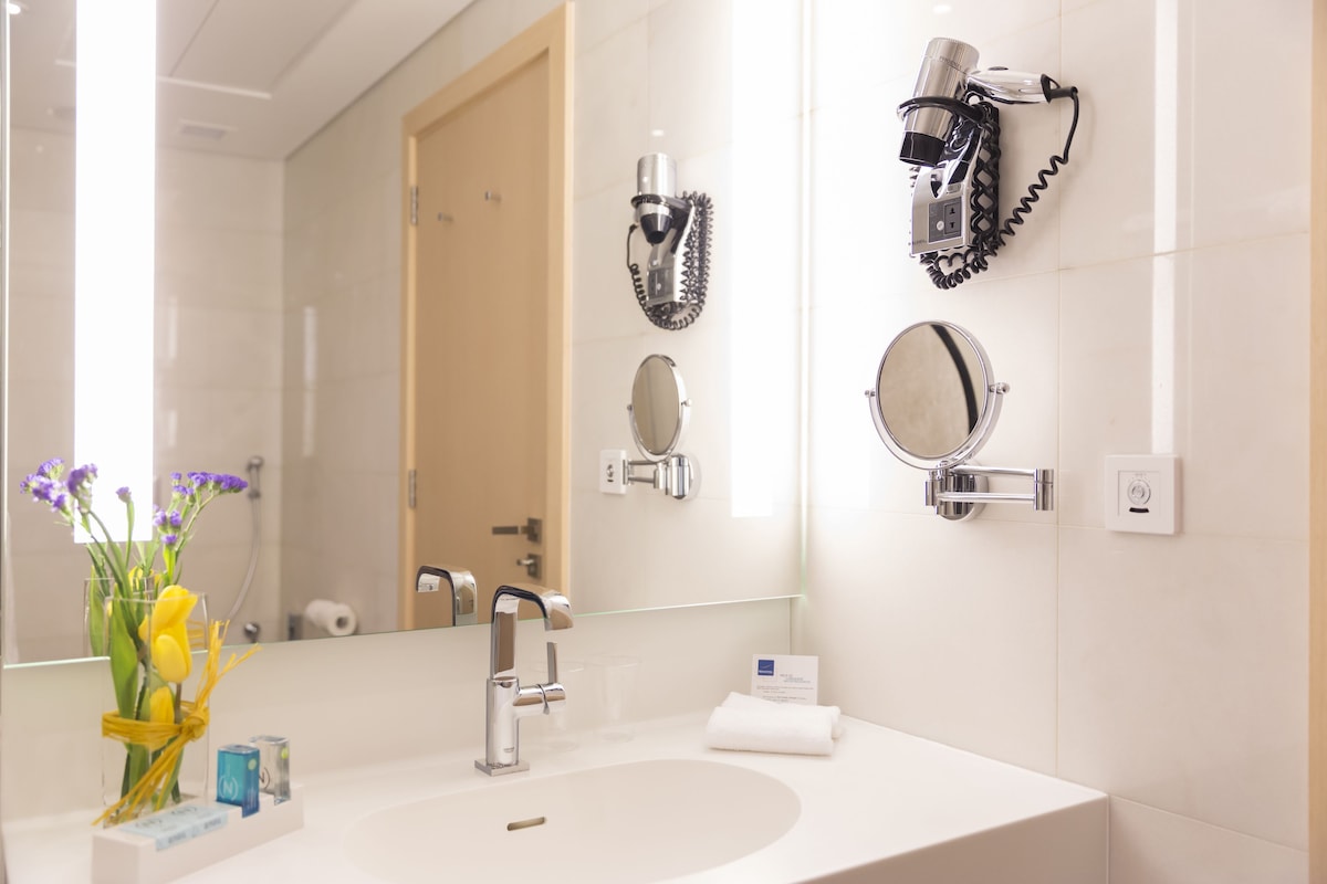 Une salle de bain d’hôtel avec un grand miroir, un lavabo et deux sèche-cheveux fixés au mur.