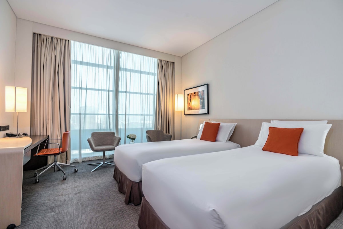 Une chambre d’hôtel avec deux lits, un bureau, une chaise et une grande fenêtre.