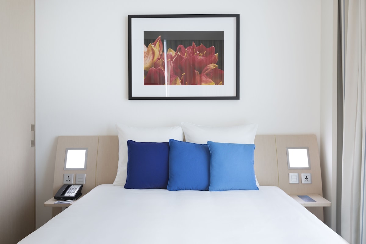 Une chambre d’hôtel avec un grand lit, deux tables de chevet, un tableau encadré au mur et une fenêtre avec des rideaux.