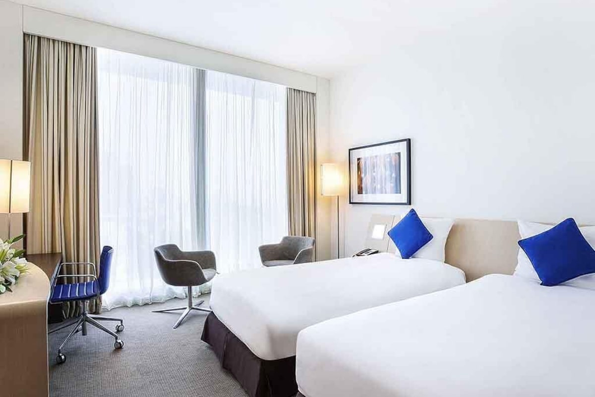 Une chambre d’hôtel avec deux lits, un bureau avec une chaise, une lampe et une grande fenêtre avec des rideaux.