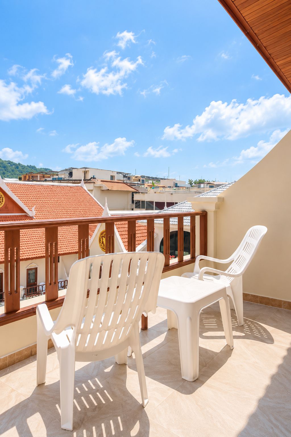 A balcony with two white plastic chairs and a table, overlooking a residential area with red-tiled roofs.