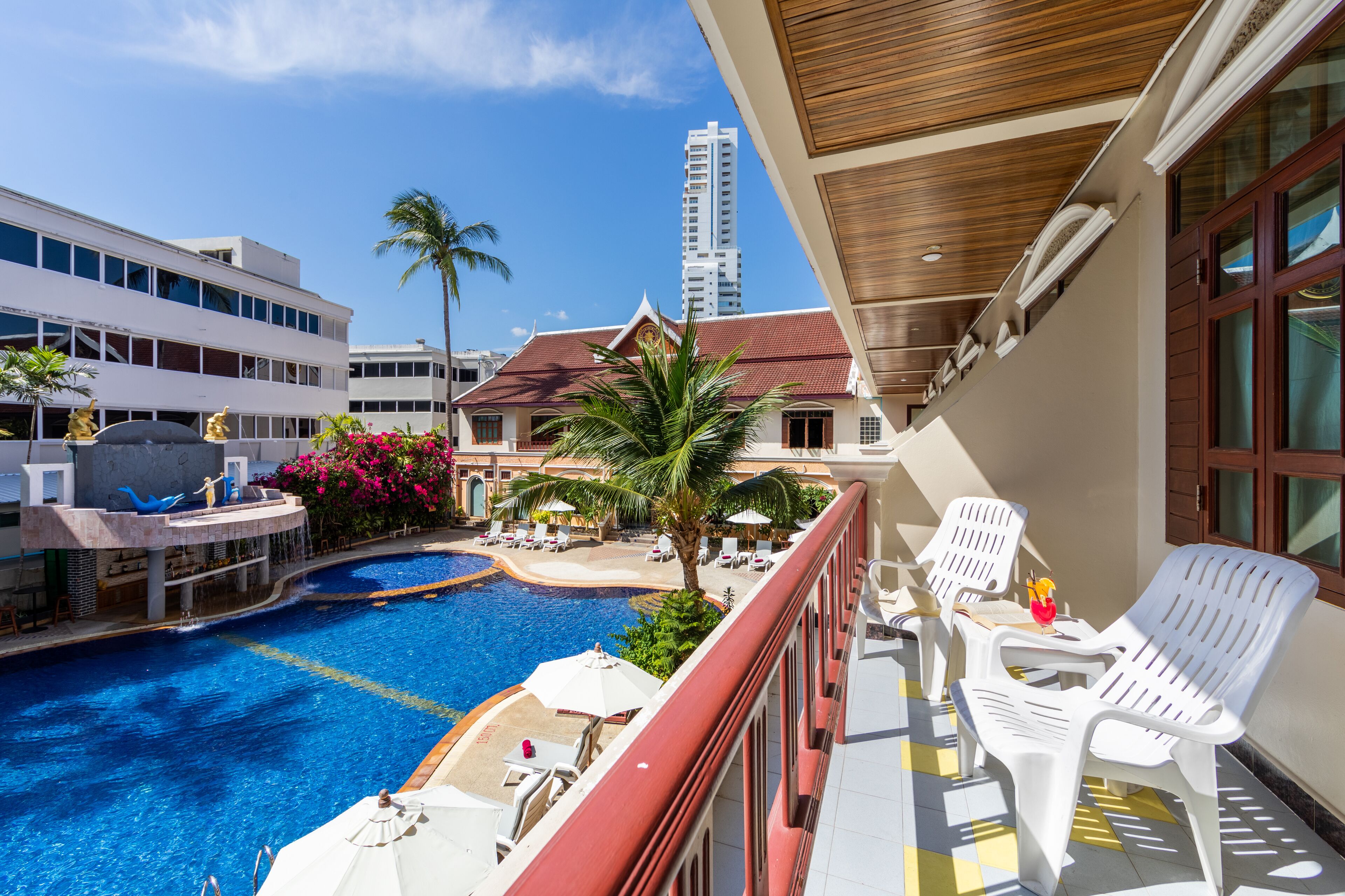 Un espace piscine d’hôtel avec vue sur un grand immeuble en arrière-plan, un plafond en bois et des chaises longues blanches.