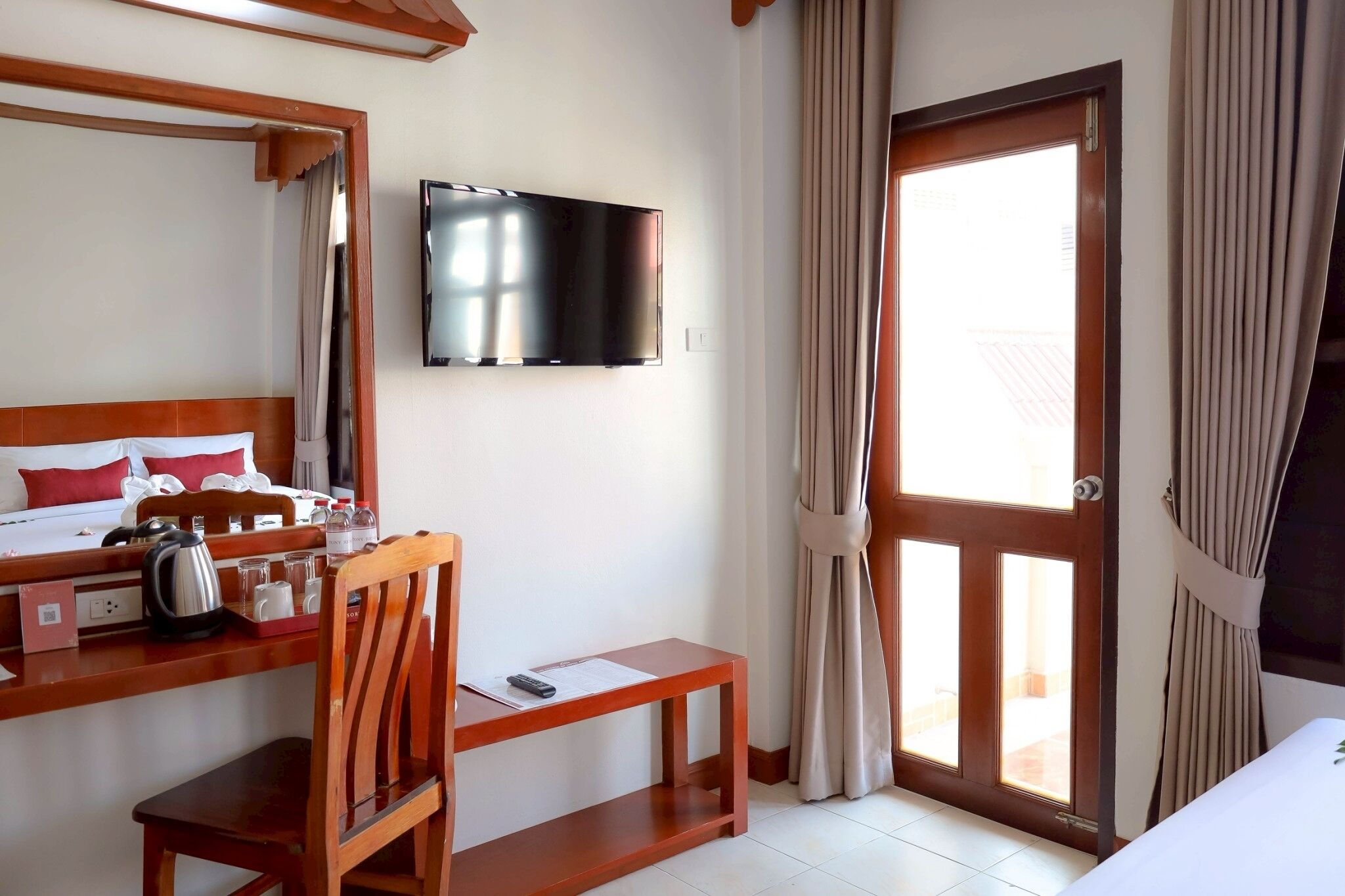 A hotel room with a bed, a desk with a kettle, a chair, a TV mounted on the wall, and a sliding glass door to the outside.